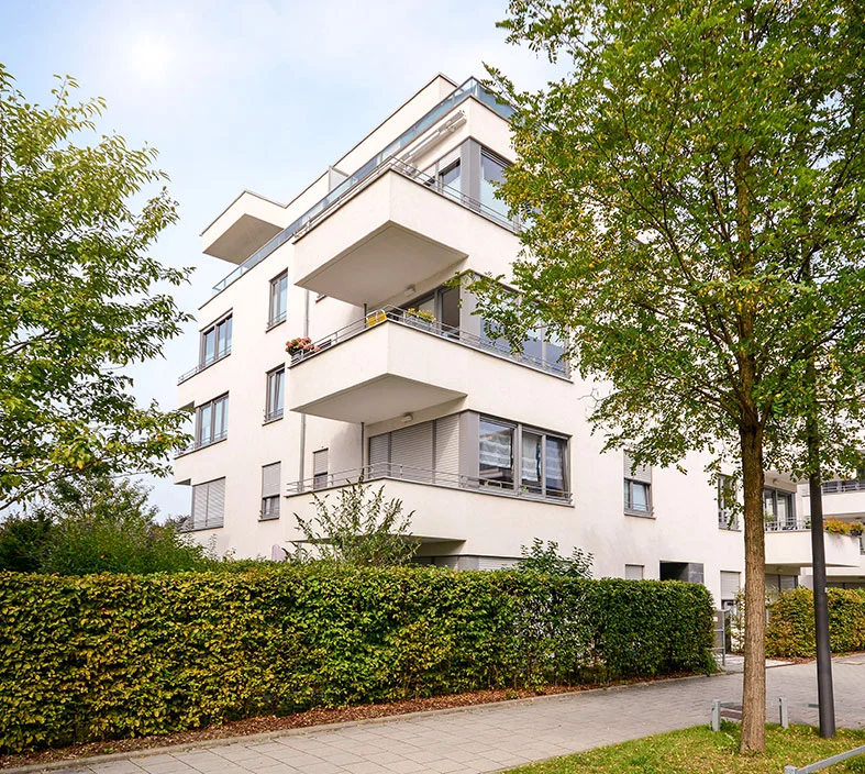 White apartment building partially shaded by nearby trees and hedges, illustrating potential obstructions to solar panel sunlight exposure.