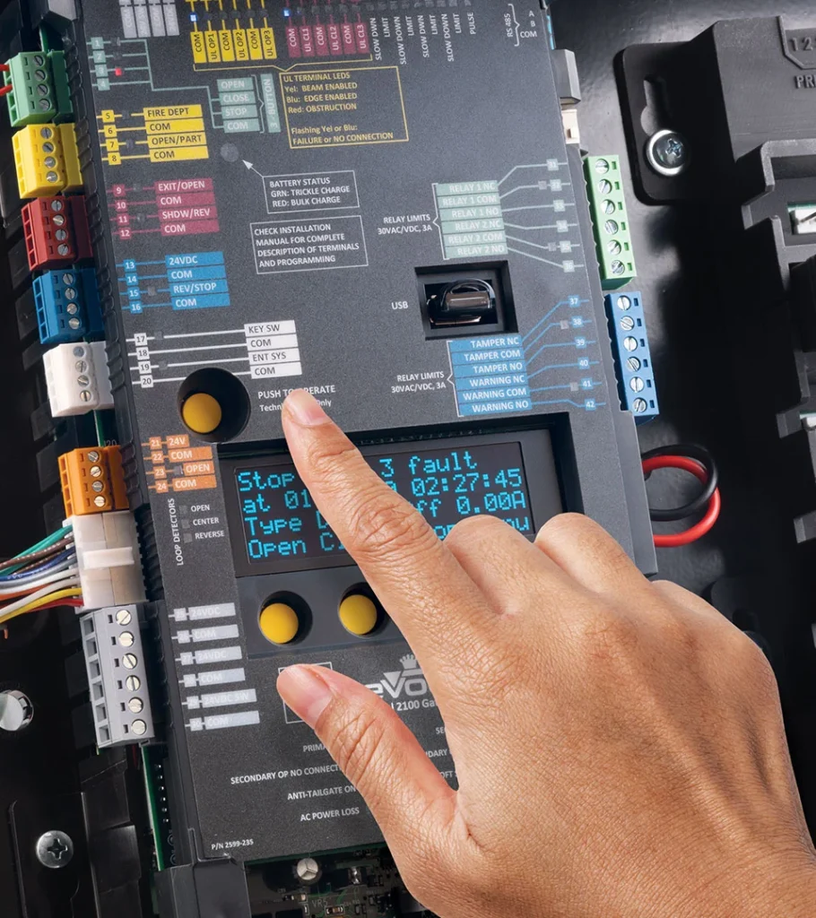 Hand interacting with the DKS 2100 controller board inside an eVolve gate operator, showing diagnostic display, wiring terminals, and system programming interface.